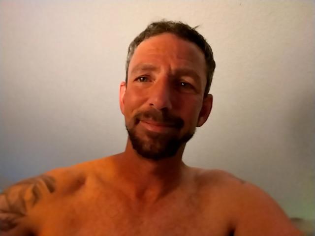 TreemanDude, shirtless with a tattoo on his right shoulder, has a slight smile, short brown hair, and a beard. His chest is hairy. He's looking at the camera.