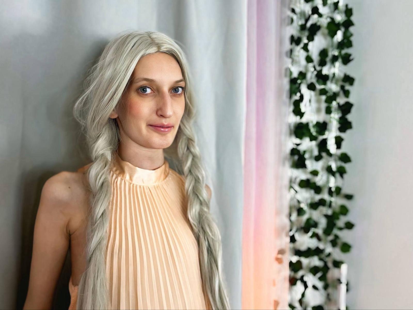 ViktoriaClarrk with long, wavy, silver hair, wearing a beige, pleated top, standing with a slight smile, and a small nose, in a well-lit, indoor setting.