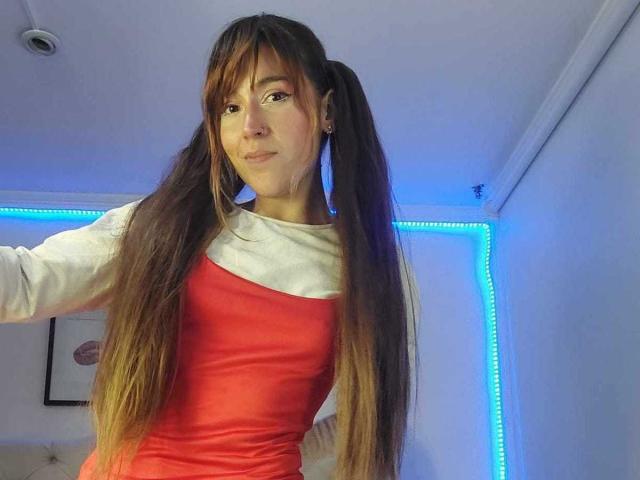 AmyCorinas with long brown pigtails, wearing a red dress with white sleeves, smiling slightly, blue LED strip lights in the background, and a small framed picture on the wall.