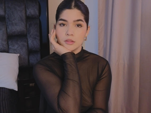 ScarlletZhen with dark hair, wearing a sheer black top, resting her cheek on her hand, looking serious. Her tits are subtly visible through the fabric.