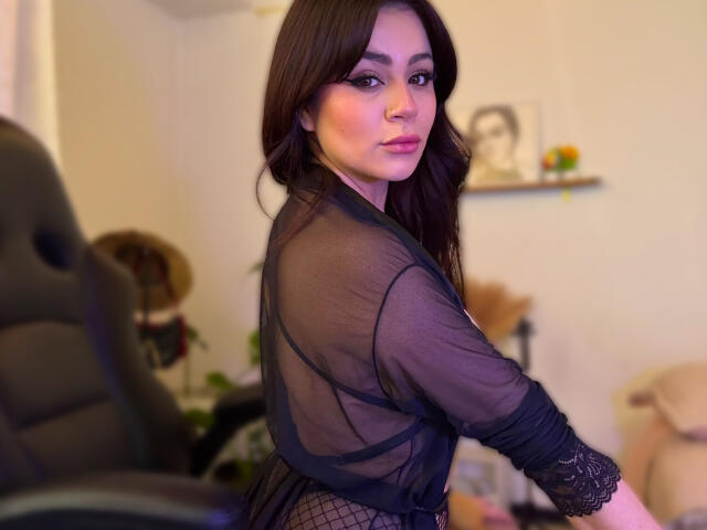 ShantallParis with long dark hair, wearing a sheer black lace top, looking over her shoulder, showing her ass and fishnet bottom, in a softly lit room.