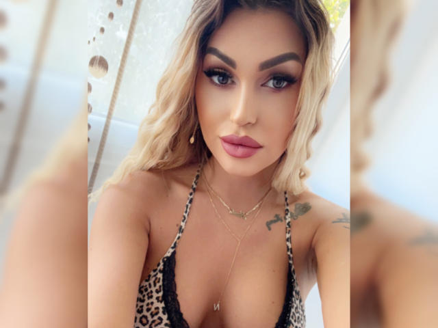 NicolSNaya has blonde hair, heavy makeup, and pink lips, wearing a leopard print bra that shows her big tits, with tattoos on her right arm, and a necklace.