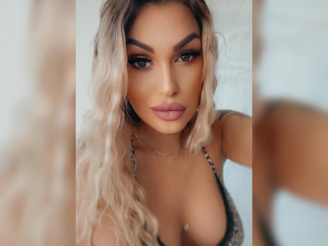 NicolSNaya hat blondes, welliges Haar, dunkle Augenbrauen, volle, rosafarbene Lippen und große Brüste, trägt ein graues Oberteil und eine goldene Halskette und blickt in die Kamera..