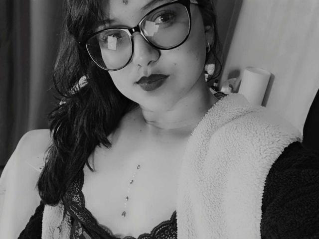 Black-and-white selfie of AzulMoon with glasses, dark lipstick, lace bra, chest with nipple piercings, and a fluffy robe, showing her tits and a relaxed expression.
