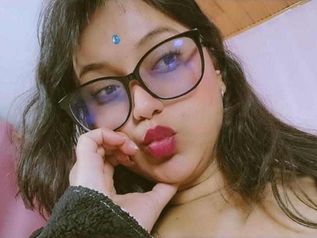 AzulMoon with big blue-tinted glasses, dark wavy hair, red lipstick, and a blue gem on her forehead, pouting with her hand near her face. She's wearing a fuzzy black sweater.