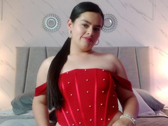 ValeriaNoir, Latina with long black ponytail, wearing a red off-shoulder dress with pearl studs, pearl bracelet, and earrings, standing in front of a gray headboard.
