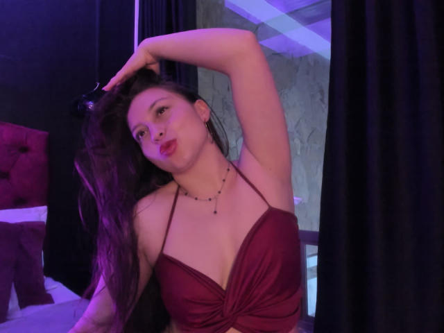 WidnyBeaumont con lunghi capelli scuri, vestito rosso e collana, con i capelli raccolti, espressione accigliata, seni visibili, in una stanza illuminata debolmente di viola..