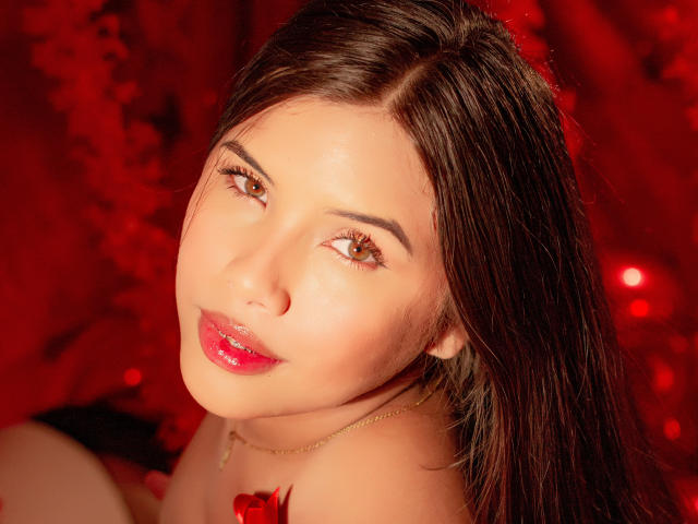 Ariy has shiny brown eyes, glossy red lips, and dark straight hair. She wears a red rose necklace against a red, bokeh-filled background. Her skin is smooth and fair.