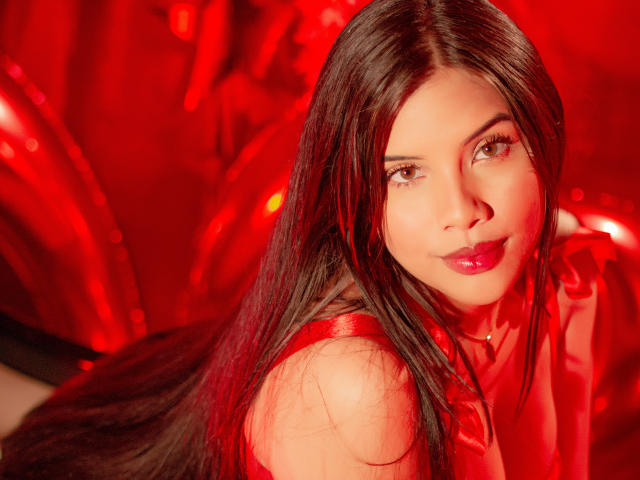 Ahriy with long dark hair, red lipstick, and a sexy smile, wearing a red top, looking over her shoulder, red light illuminating her face and chest.
