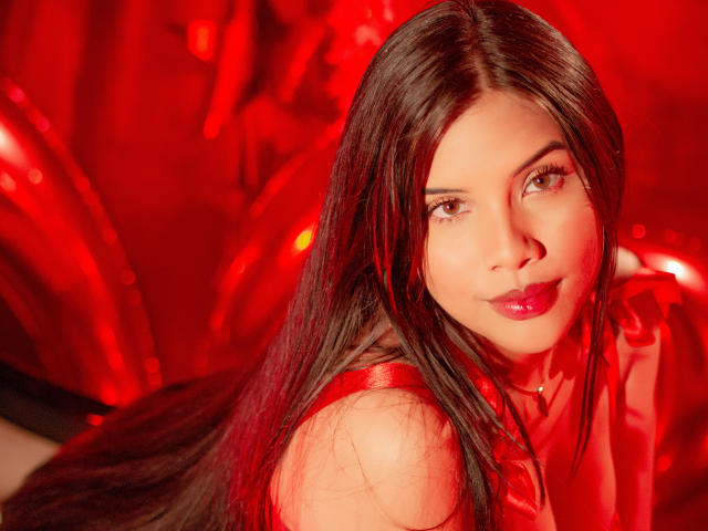Ahriy has long black hair, red lips, and brown eyes, wearing a red outfit, with a red background and red lighting, looking over her shoulder with a slight smile.