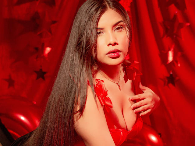 Ahriy with long black hair, red lipstick, and a red dress with ruffled straps, showing her tits, hand on chest, red background with stars, looking at camera.