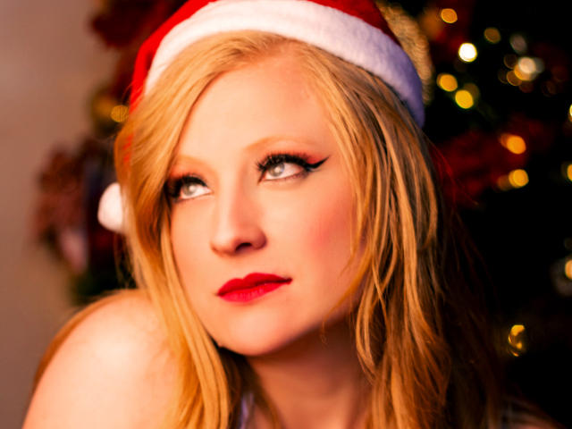 Close-up of AliceCathyHot, a blonde woman with fair skin and blue eyes, wearing a red Santa hat. Her wavy hair is loose, and she has bold red lipstick and black eyeliner. The soft, warm lighting highlights her facial features. The blurred background has bokeh lights, suggesting a festive, Christmas setting. The image focuses on AliceCathyHot's face, showing her calm, slightly curious expression. The composition centers her face, with the Santa hat adding a holiday element.