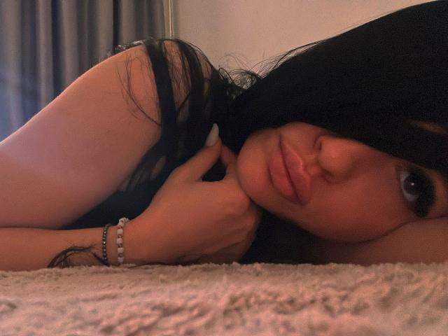 SashaLorenz, with black hair, lies on a carpet, her face near a pillow, looking at the camera, showing her tits and a bracelet on her wrist.