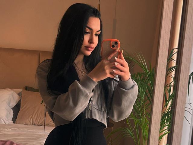SashaLorenz with long black hair, wearing a grey and black top, taking a mirror selfie with an orange phone, her ass in black pants, a potted plant behind her.