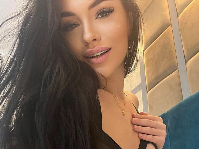 SashaLorenz with long black hair, blue eyes, and glossy pink lips, wearing a black top, holding her collar, gold necklace, slight smile, and a sexy, confident look.