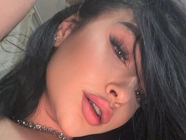 SashaLorenz has dark, wavy hair, full pink lips, and is wearing a bejeweled necklace. Her thick eyelashes and slightly parted lips are in the foreground.