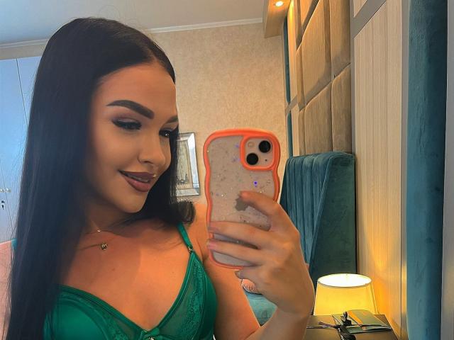 SashaLorenz with long black hair, green lace bra, holding orange phone, smiling, looking down, bedroom with green upholstered chair, white lamp, and beige wall.