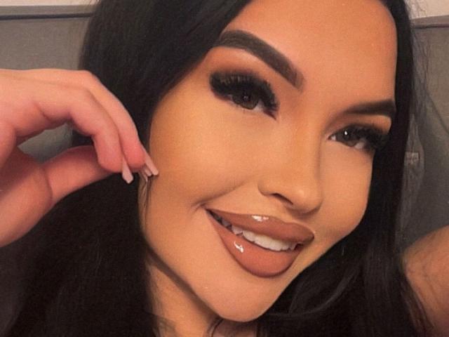 SashaLorenz has long black hair, thick eyebrows, long eyelashes, glossy brown lips, and is playfully sticking out her shiny tongue. Her hand is near her face.