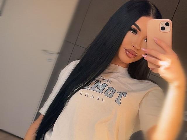 SashaLorenz with long black hair, full lips, and a white tee, taking a selfie with a pink iPhone. Boobs are partially visible. Her nails are long and pink.