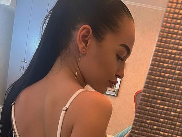 SashaLorenz with long black hair in a ponytail, white strap dress, gold earrings, and turned profile, showing her smooth back and subtle makeup in a softly lit room.