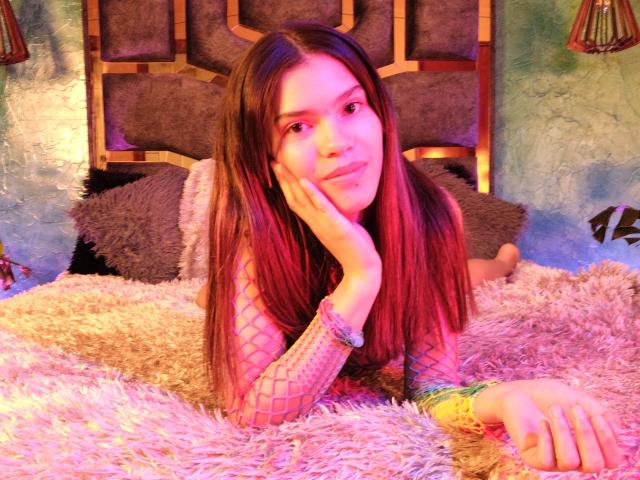 EmyCarther with pinkish-red hair, lying on a fluffy pink bed, wearing fishnet sleeves, and colorful bracelets, resting her cheek on her hand, looking at the camera.