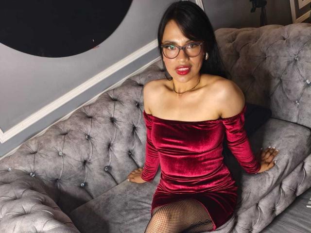 AnyJoness with red velvet off-shoulder dress, black fishnet stockings, and glasses, sitting on a tufted gray couch, showing her bare ass and medium tits.