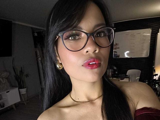 AnyJoness has shiny black hair, large glasses, glossy pink lips, and a gold necklace, with bare shoulders showing. She's wearing gold earrings and a red off-shoulder top.