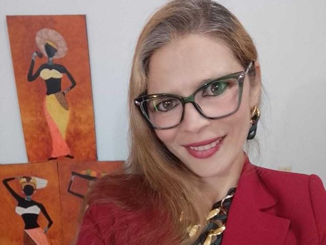ParisMatur69 with long blonde hair, green glasses, red lipstick, red blazer, gold necklace, smiling at camera, standing next to colorful African-style paintings.