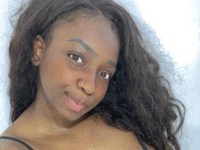 AphroditeSass has dark brown skin, curly black hair, a nose ring, and a slight smile with pink lips. She's wearing a black strap top.