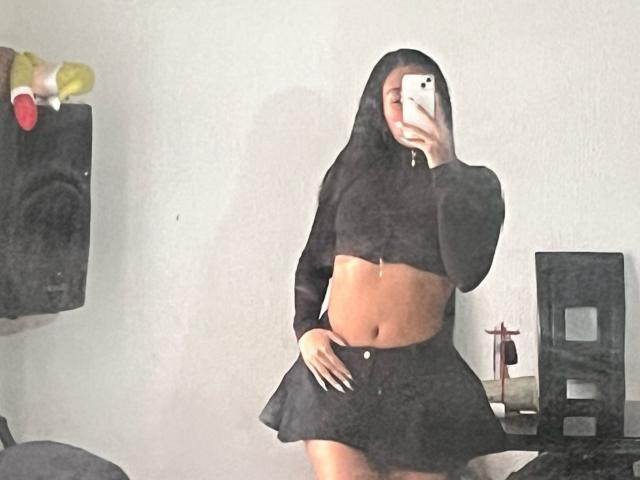 ElaStone mit mittelbrauner Haut, langen schwarzen Haaren unter einer Kapuze, einem schwarzen Crop-Top und einem Rock, hält ein Telefon, zeigt einen flachen Bauch und einen kleinen Hintern und steht in einem Raum..