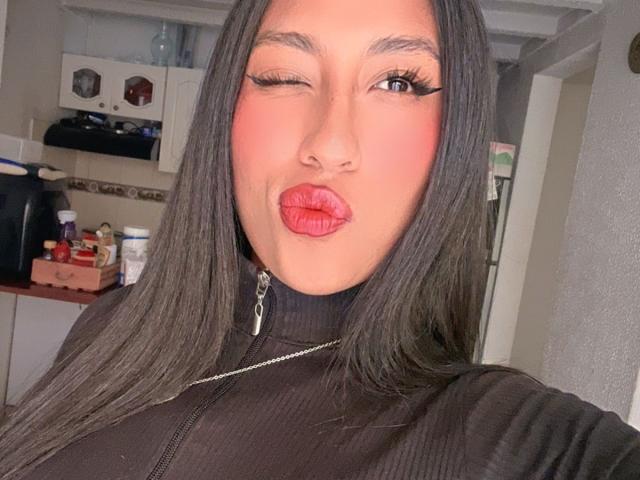 ElaStone, knipend met haar ogen, met lang zwart haar, een zwarte ritsjas aan, volle rode lippen en zwarte eyeliner, maakt een kusgezicht. Haar borsten zijn bedekt door de ritsjas..