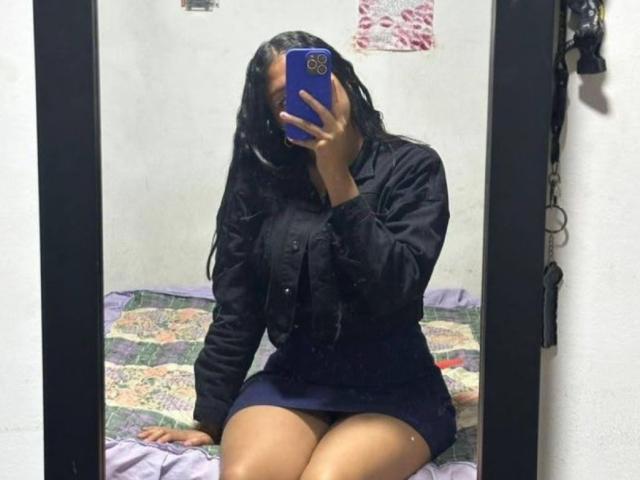 ElaStone, com longos cabelos pretos, vestindo uma camisa preta e uma saia azul curta, está sentada em uma cama, segurando um telefone azul, cobrindo o rosto, mostrando suas nádegas..