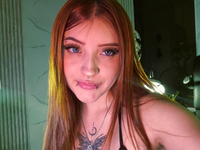 MaylinSanchez with red hair, blue eyes, and multiple facial piercings, including a nose ring and lip ring, wearing a black top, showing her chest tattoo.