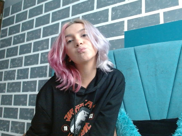 AlterEgoSecond with pink and white hair, puffy black hoodie, and a pouty face, sitting on a blue, tufted headboard. Soft pink hair, cute pout.