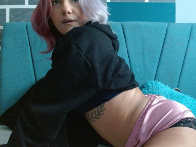 AlterEgoSecond with pink and white hair, black hoodie, pink satin shorts, black lace, and a leaf tattoo, leaning on a teal couch, showing her ass and looking at camera.