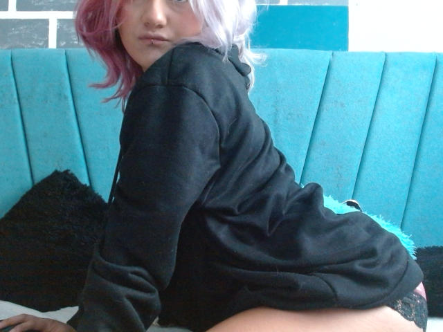 AlterEgoSecond with pink hair, black hoodie, and black lace panties, showing ass, kneeling on bed, looking over shoulder, blue upholstered headboard in background.