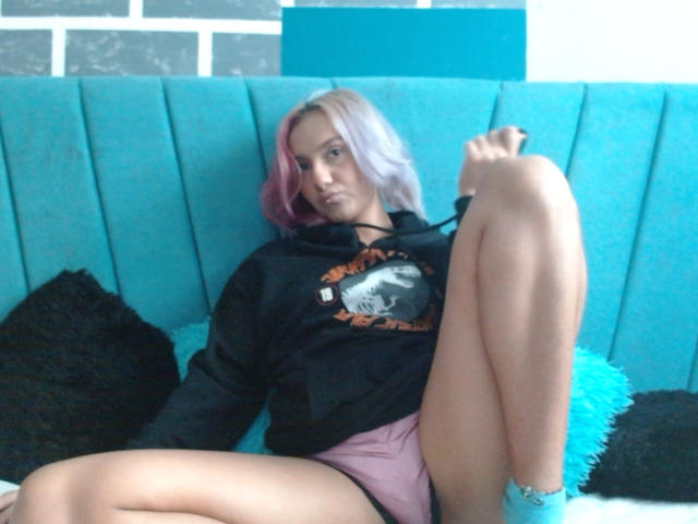 AlterEgoSecond with pink hair, black hoodie, and pink shorts, legs spread, foot up, blue socks, sitting on turquoise couch, looking at camera, slightly annoyed expression.