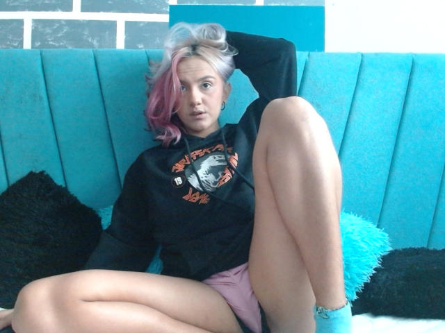 AlterEgoSecond with pink and white hair, black hoodie, pink shorts, and blue socks, legs spread, sitting on teal couch, looking at camera, hand behind head.