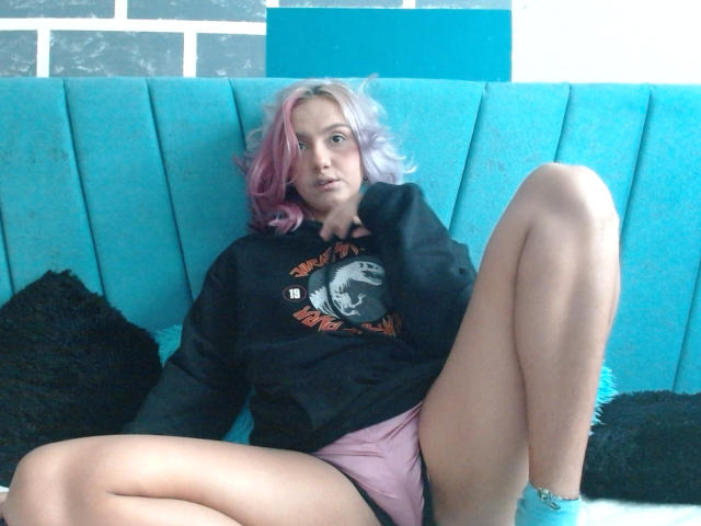 AlterEgoSecond with pink and purple hair, wearing a black hoodie and pink shorts, sitting on a blue couch with legs spread, looking at the camera with a neutral expression.