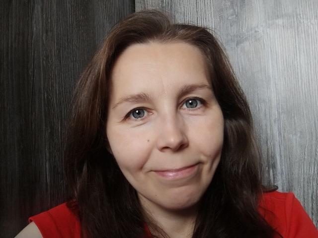 DaisyEdit has medium-length brown hair, slightly wavy, with a few strands falling over her face. Her blue eyes are slightly upturned, giving a subtle, friendly look. She has a small mole on her right cheek and a slight, closed-lip smile. She's wearing a bright red top, which contrasts with her fair skin. The texture of her hair is soft and natural, while the background is a dark, wood-like pattern. The image is a close-up, focusing on her face, with even lighting that highlights her features.