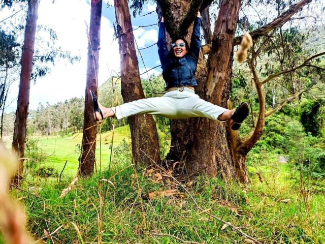JessyLuxs hangs from a tree with arms up, legs spread, wearing white pants and a blue jacket, smiling with sunglasses on, surrounded by greenery.