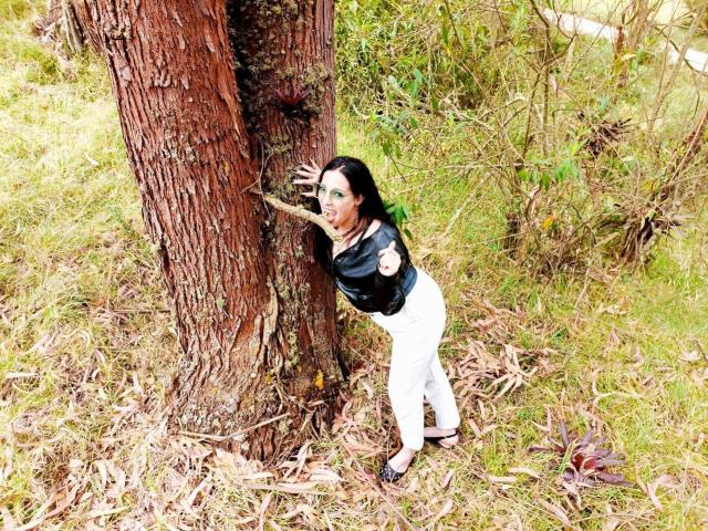 JessyLuxs leaning against a tree, wearing a black leather top and white pants, with black flats, and her hand on the trunk, in a grassy outdoor setting.