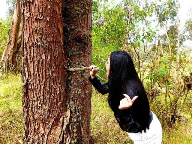 JessyLuxs with long black hair, black top, and white skirt, sticking a twig into a tree's rough, brown bark, right hand raised, standing in a grassy forest.
