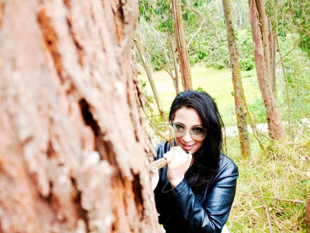 JessyLuxs with black hair, glasses, and leather jacket, smiling and biting her finger, standing next to a large, textured tree trunk.