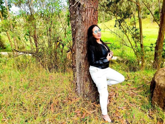 JessyLuxs leaning against a tree, black leather jacket, white pants, long black hair, green glasses, smiling, holding something in her hand, standing in a grassy forest.