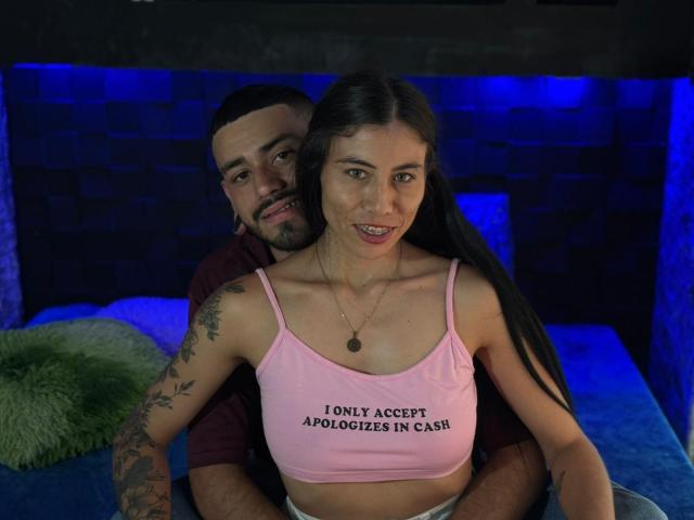 TashaAndRolo with long black hair, pink crop top, small tits, tattoos, and necklace, sitting on bed, Rolo behind her, smiling, blue-lit background, green pillow on left.
