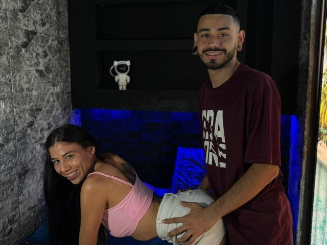TashaAndRolo, a slim woman with long black hair and small tits, bends over while Rolo, a cute man with a beard, grabs her white butt in a pink top.