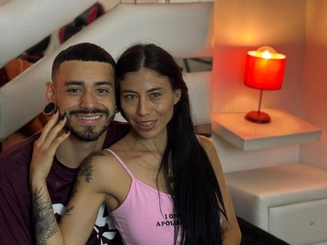 TashaAndRolo, a tan-skinned dude with a beard and black ear gauges, and a tan-skinned woman with long black hair, smiling and touching cheeks in a cozy room.