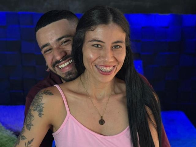 TashaAndRolo with long black hair and pink top, smiling, has tattoos on her right arm. Rolo, with short black hair and beard, stands behind her, grinning.