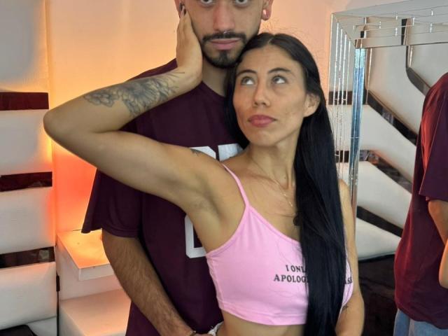 TashaAndRolo with long black hair, pink sports bra, and small tits, stands in front of Rolo, who has a tattooed arm, holding her chin. Rolo wears a maroon shirt.